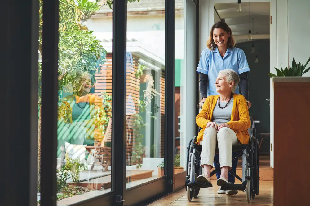 a senior woman sitting in her wheelchair by a large window with her caregiver